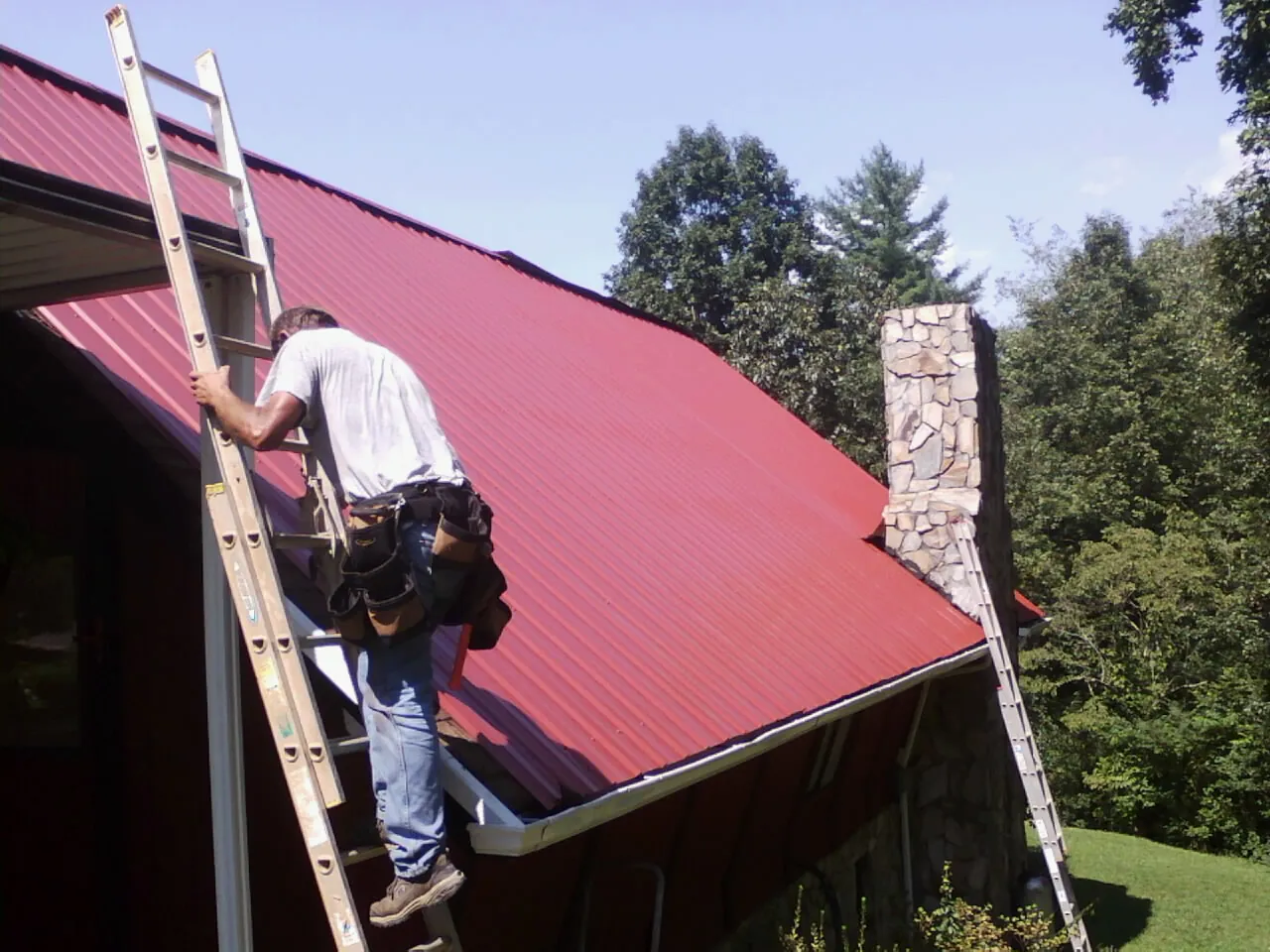 Roofer on a ladder beside a red metal roof and stone chimney — Soffit & Fascia Repair by Roofing Brothers in Bolivar